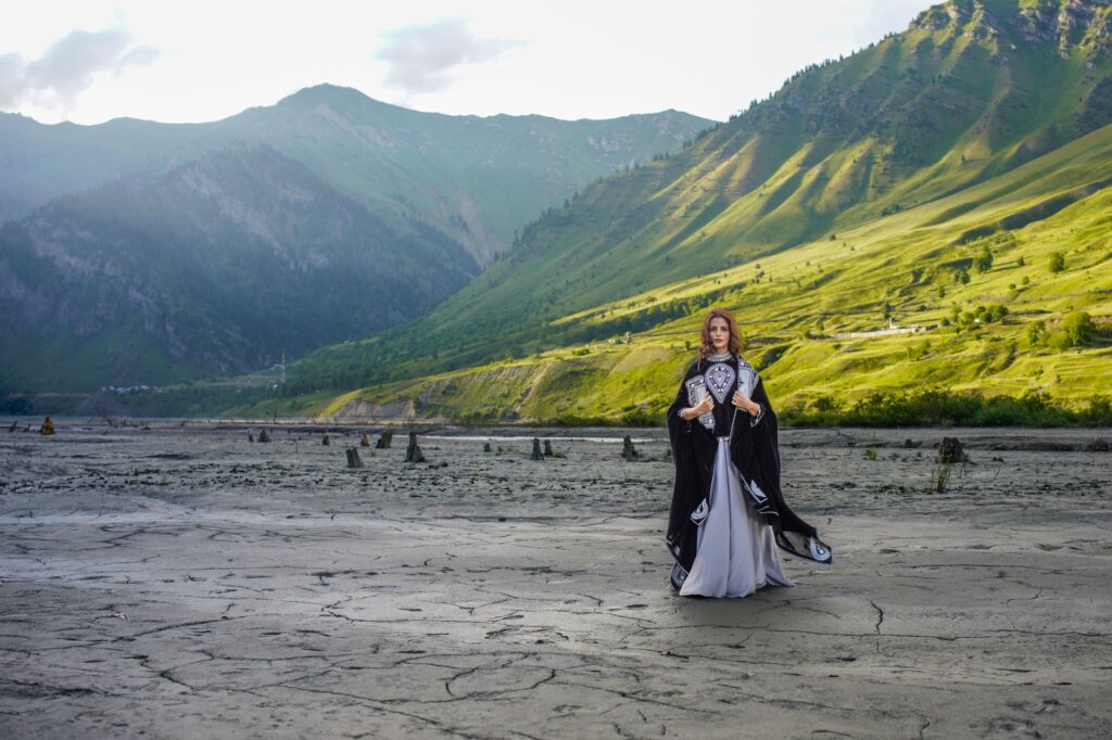 A woman in an elegant costume stands amidst a stunning mountainous landscape during the day.