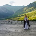 A woman in an elegant costume stands amidst a stunning mountainous landscape during the day.