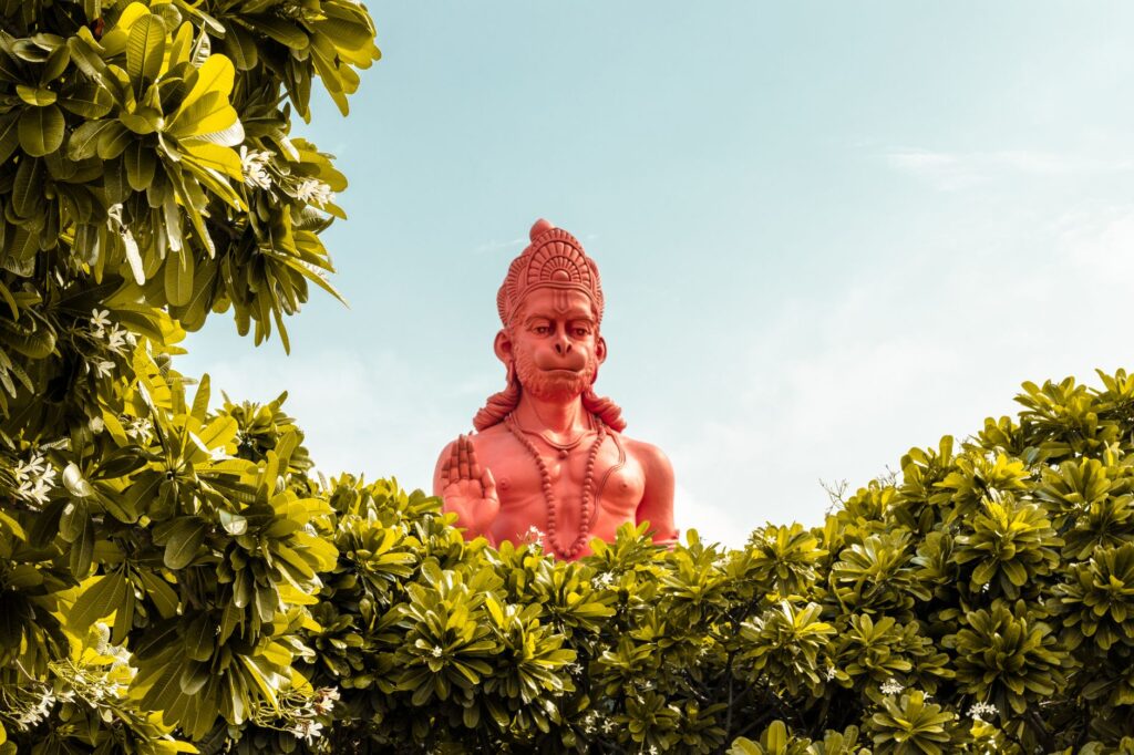 A vibrant red statue of Hanuman surrounded by green foliage under a clear sky.