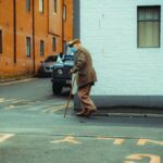 Senior man with cane walking past brick building on urban sidewalk.
