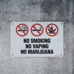 A clear warning sign prohibiting smoking, vaping, and marijuana on a concrete wall.
