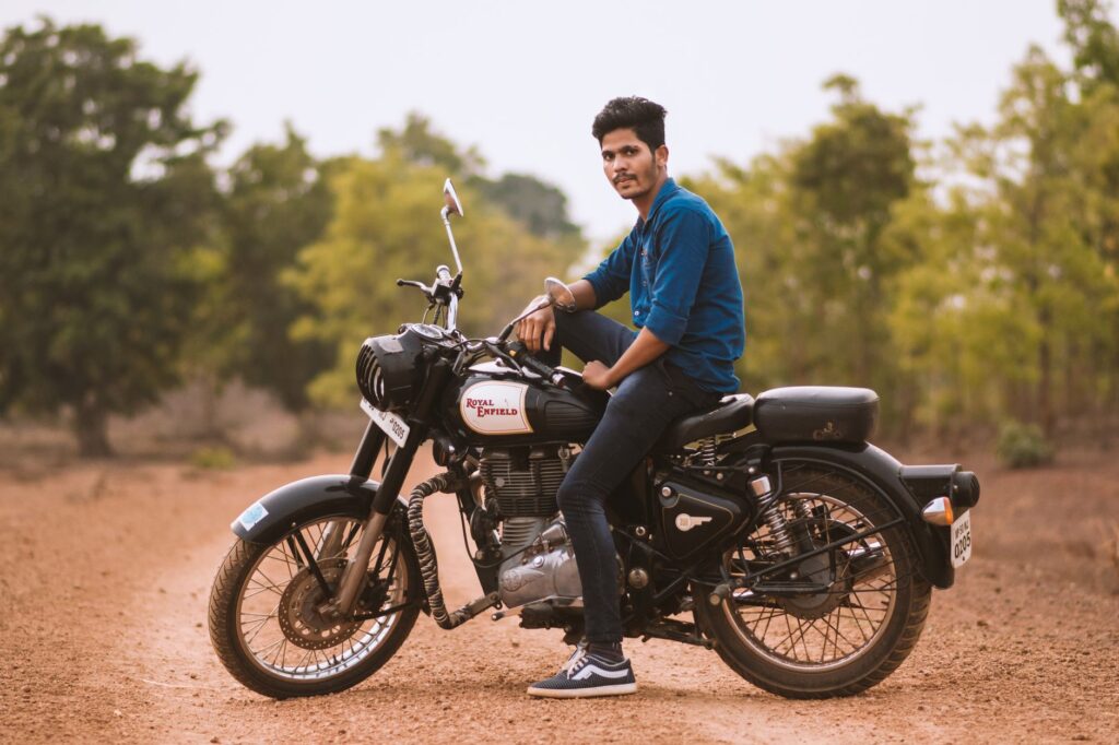 A young South Asian man sitting on a Royal Enfield motorcycle in an outdoor rural setting.