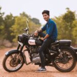 A young South Asian man sitting on a Royal Enfield motorcycle in an outdoor rural setting.