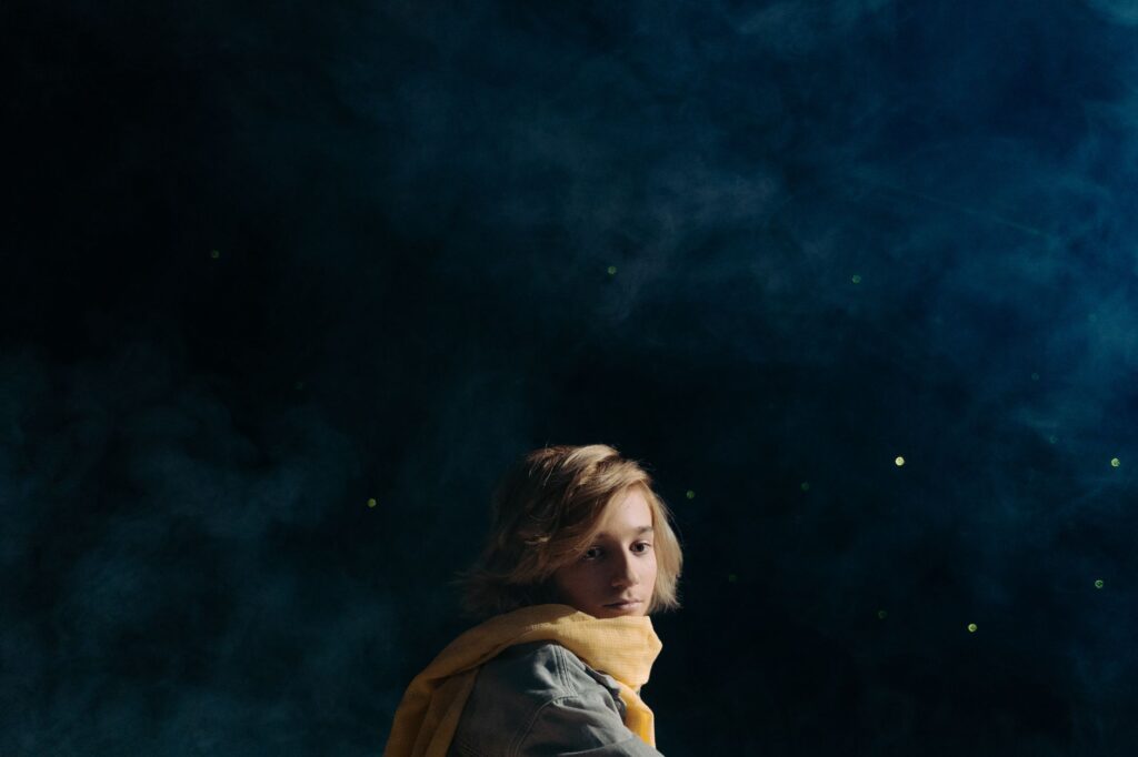 A contemplative young boy with a yellow scarf sits against a starry night sky backdrop, embodying a literary character.