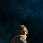 A contemplative young boy with a yellow scarf sits against a starry night sky backdrop, embodying a literary character.