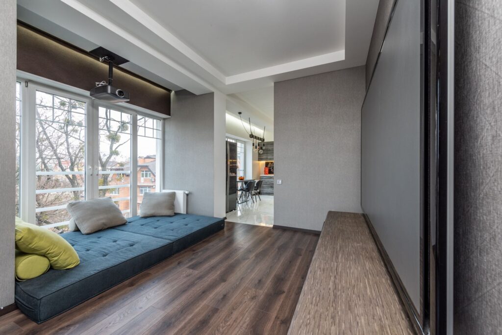 Blue soft mattress with colorful cushions near window against wall with modern TV in spacious apartment with projector and kitchen