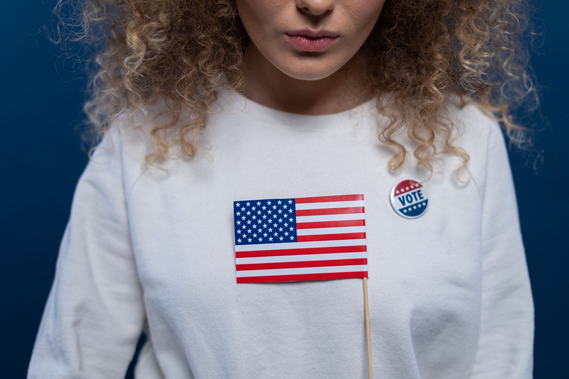A woman with curly hair holding an American flag and wearing a vote pin against a blue background.