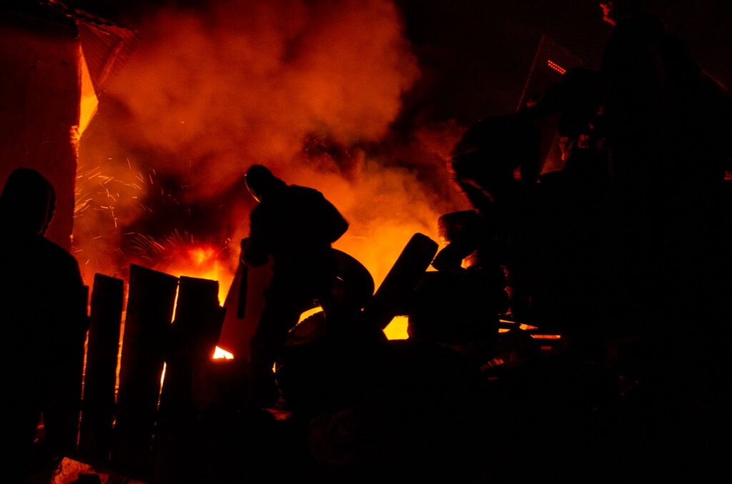 Silhouetted people stand near a blazing fire, creating dramatic shadows and vibrant colors.