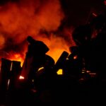 Silhouetted people stand near a blazing fire, creating dramatic shadows and vibrant colors.