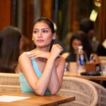 A young woman in a turquoise top sits in a lively, crowded restaurant, appearing thoughtful.