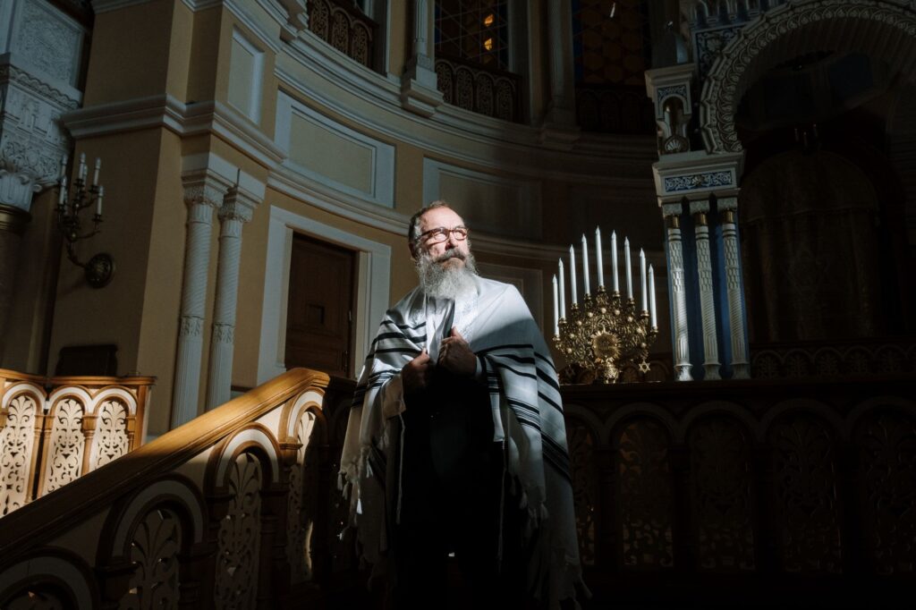 A bearded rabbi stands in a richly decorated synagogue, holding a tallit.