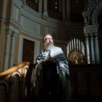 A bearded rabbi stands in a richly decorated synagogue, holding a tallit.