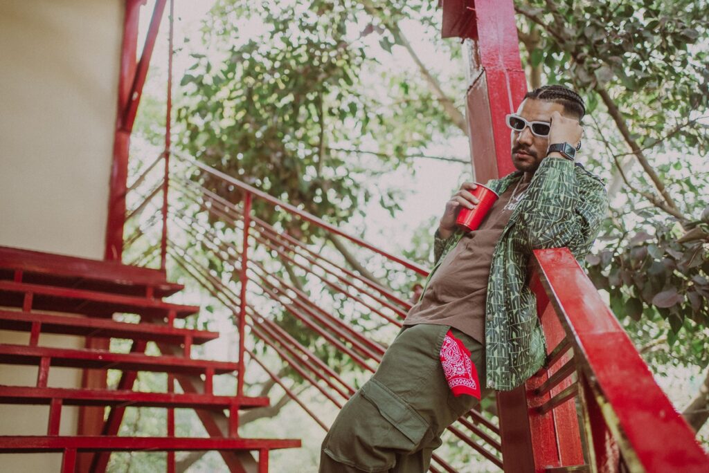 Fashionable man with braided hair and sunglasses holding a red cup, leaning on a staircase outdoors.