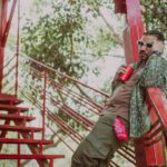 Fashionable man with braided hair and sunglasses holding a red cup, leaning on a staircase outdoors.