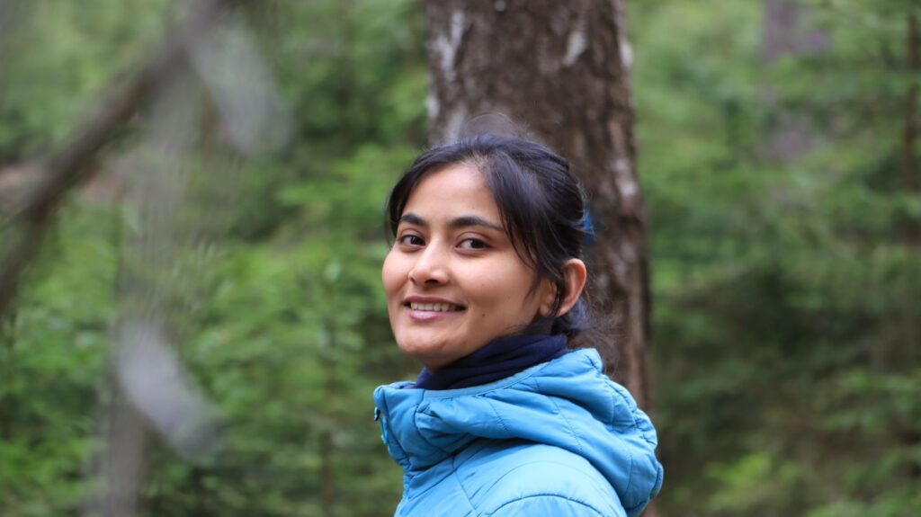 Portrait of a smiling woman in a blue jacket amidst a lush green forest.