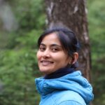 Portrait of a smiling woman in a blue jacket amidst a lush green forest.