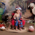 Adorable child dressed as Krishna, sitting with peacock and cow in a colorful setting.