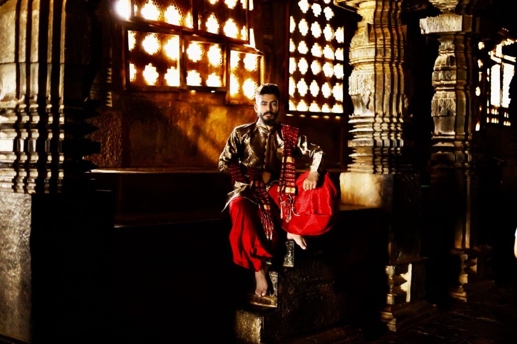 A man dressed in traditional clothing sits inside an ornately decorated temple with golden lighting.