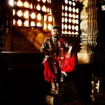 A man dressed in traditional clothing sits inside an ornately decorated temple with golden lighting.