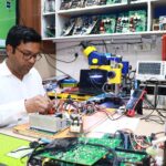 An electronics engineer working on circuit board repairs in a tech workshop filled with tools and equipment.