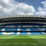 Etihad Stadium in Manchester with empty blue seats under a cloudy sky.
