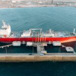 Aerial shot of a red cargo ship docked at an industrial port with clear blue water.