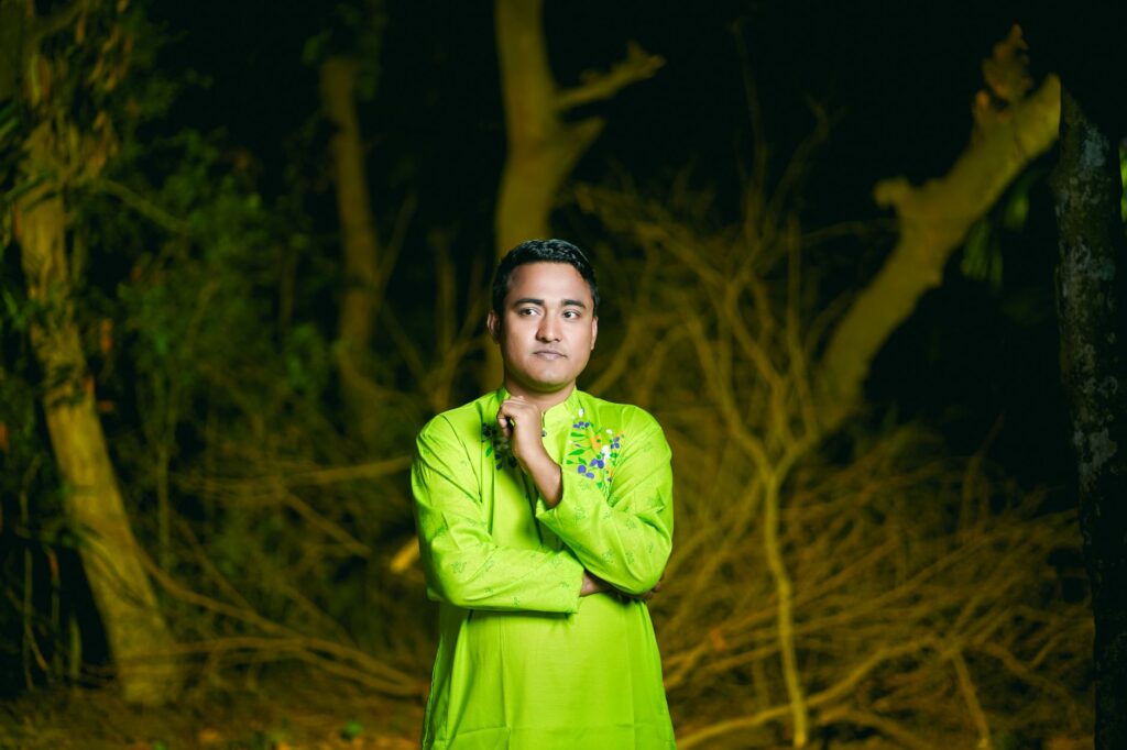 A portrait of a man in vibrant green traditional wear posing outdoors amidst trees at night.