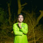A portrait of a man in vibrant green traditional wear posing outdoors amidst trees at night.