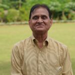 Senior man in a yellow shirt smiling outdoors in a lush green garden.