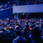 A crowded conference hall with diverse audience attentively listening to a speaker.