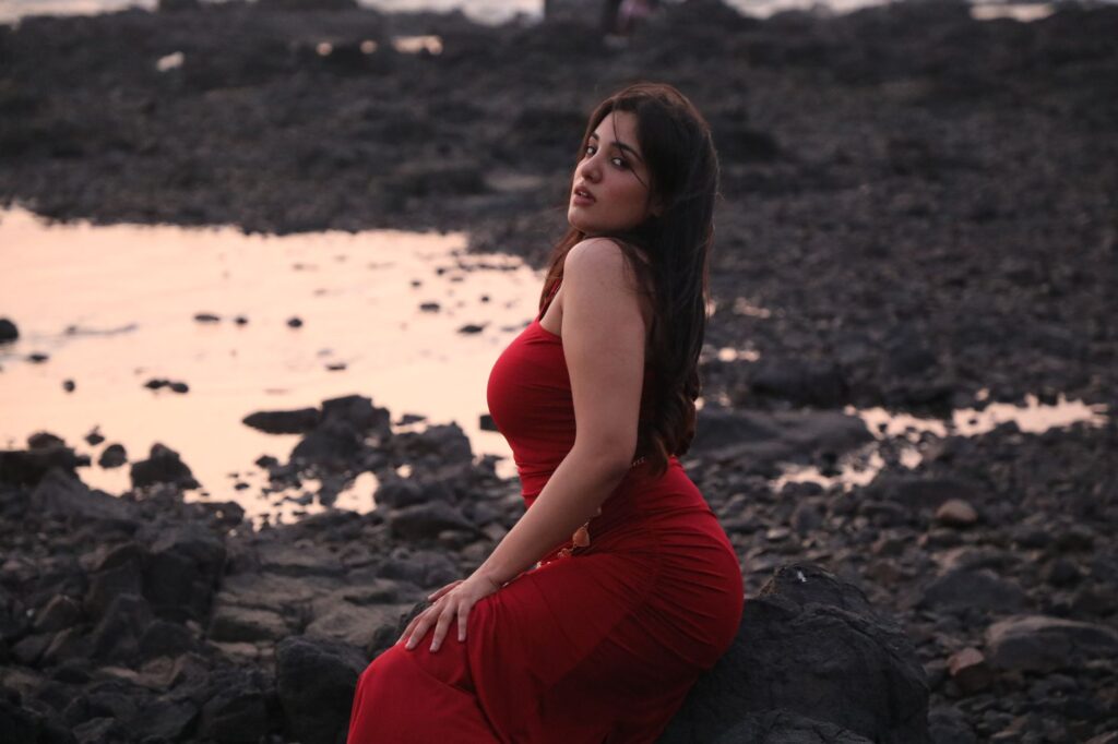 Woman in a red dress sitting on rocks by the beach during sunset, evoking elegance and tranquility.