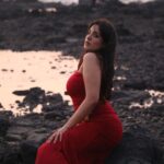 Woman in a red dress sitting on rocks by the beach during sunset, evoking elegance and tranquility.