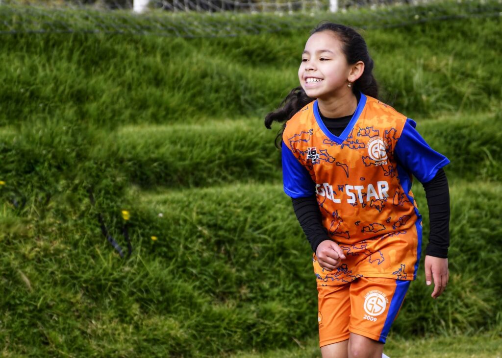 A young girl in vibrant sportswear joyfully running on a green field during soccer practice.
