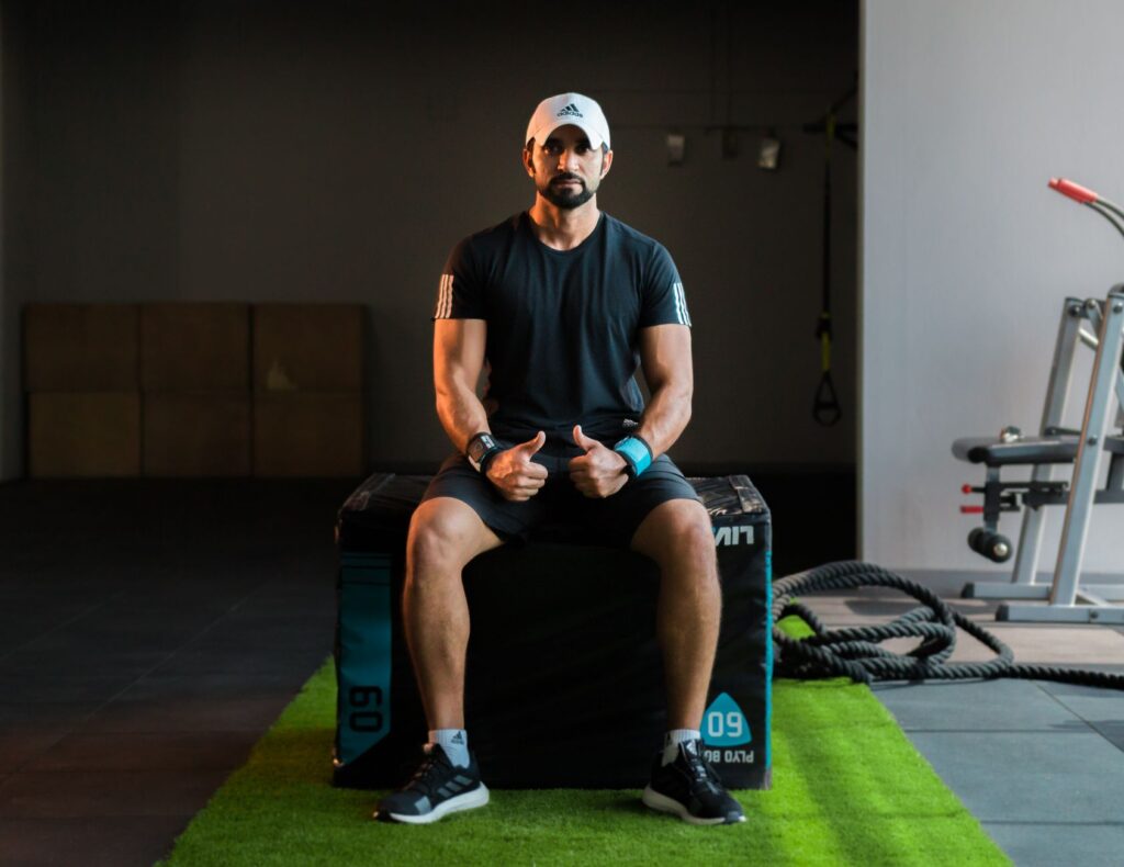 Middle Eastern adult male in gym attire sitting on plyo box, giving thumbs up.