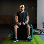 Middle Eastern adult male in gym attire sitting on plyo box, giving thumbs up.