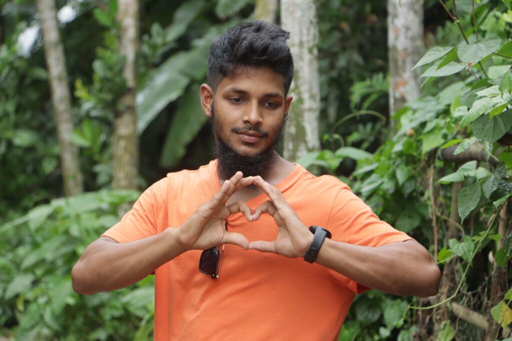 A young man in an orange shirt forms a heart gesture surrounded by vibrant greenery.