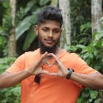 A young man in an orange shirt forms a heart gesture surrounded by vibrant greenery.