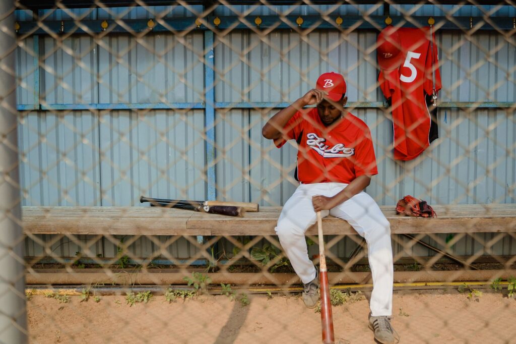 Athlete in uniform seated in a dugout bench holding a baseball bat.