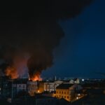 A dramatic scene of a fire with smoke rising over the cityscape of Kyiv at night.
