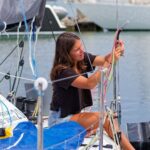 A young woman adjusts a sail on a docked sailboat, showcasing her sailing skills.
