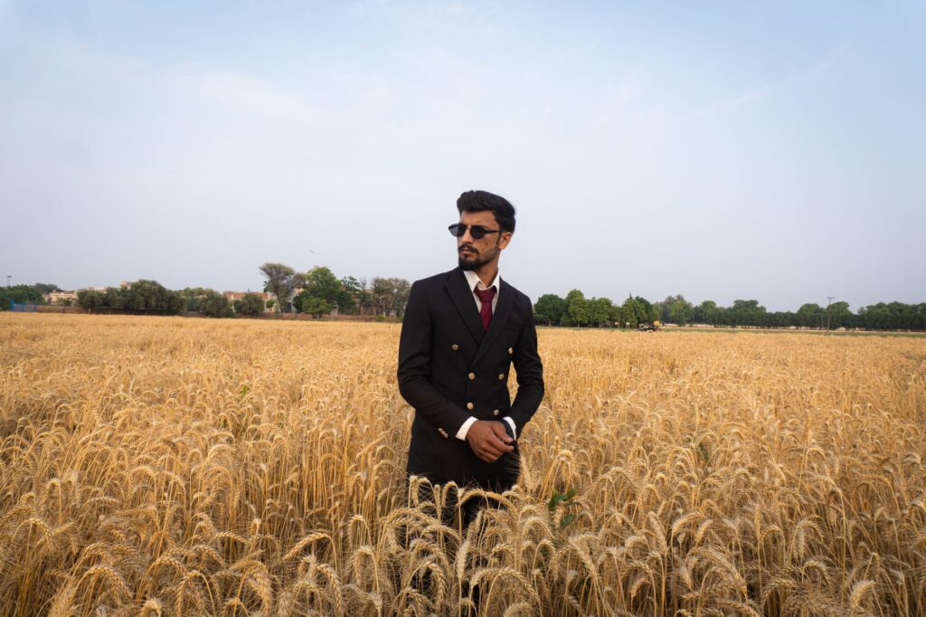 A stylish young man in a black suit stands confidently in a golden wheat field under a clear sky.