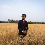 A stylish young man in a black suit stands confidently in a golden wheat field under a clear sky.