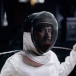 Fencer in full gear holding a foil, captured indoors with a focused expression.