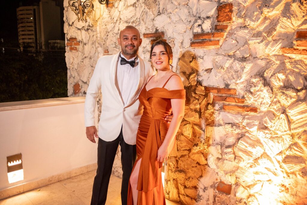 Stylish couple in formal attire poses against illuminated stone wall, showcasing evening elegance.