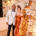 Stylish couple in formal attire poses against illuminated stone wall, showcasing evening elegance.