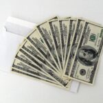 Fan of US $100 bills partially out of a white envelope on a white background.