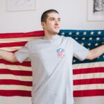 Man holds American flag indoors, expressing patriotism with casual NFL shirt.