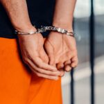 Close-up of person in handcuffs wearing orange attire behind prison bars, symbolizing arrest.