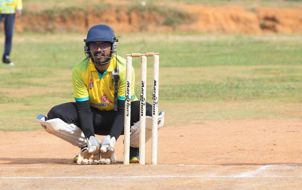 Cricket wicketkeeper crouching behind stumps during a match on a sunny day on the field.
