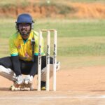 Cricket wicketkeeper crouching behind stumps during a match on a sunny day on the field.